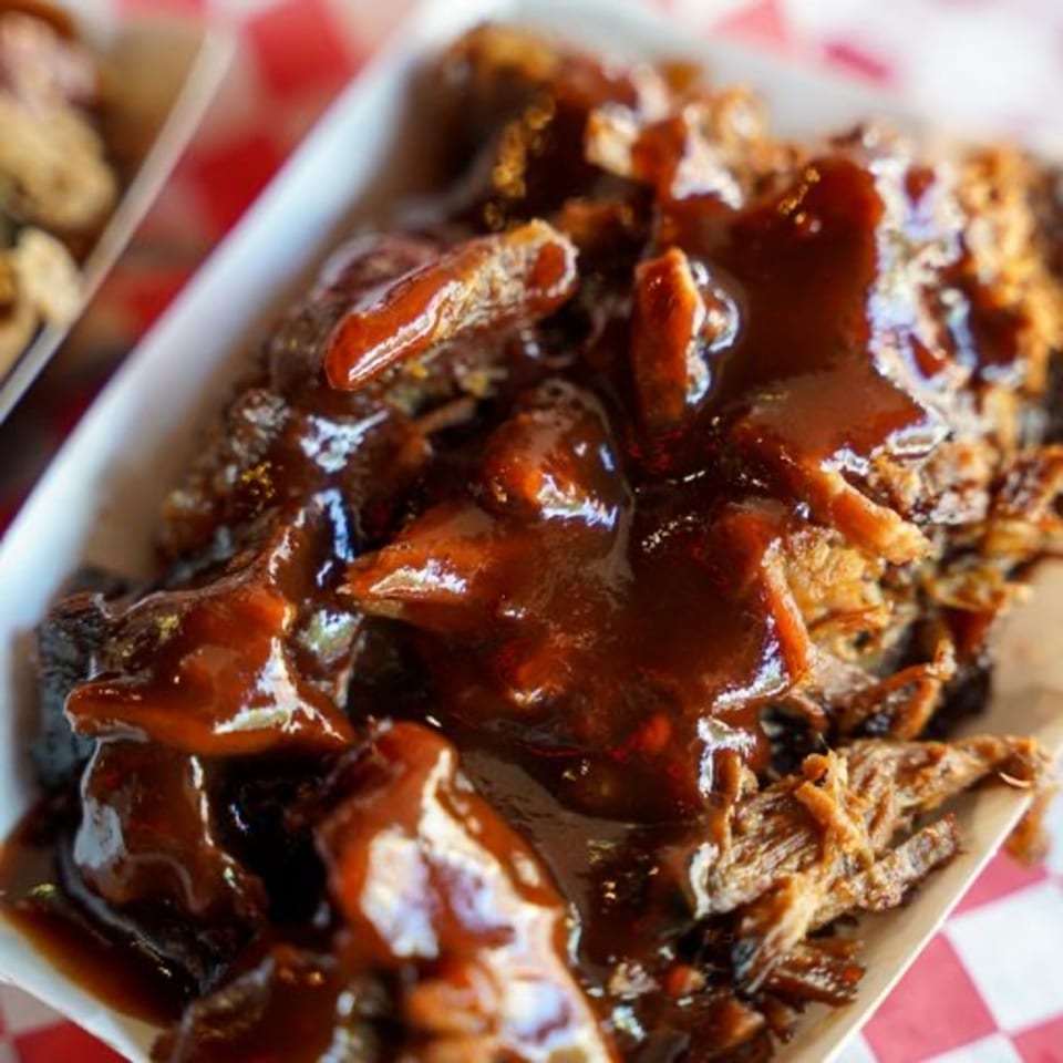 Close-up of Real Smoq’ed BBQ pulled pork covered in rich, dark barbecue sauce in a white tray.