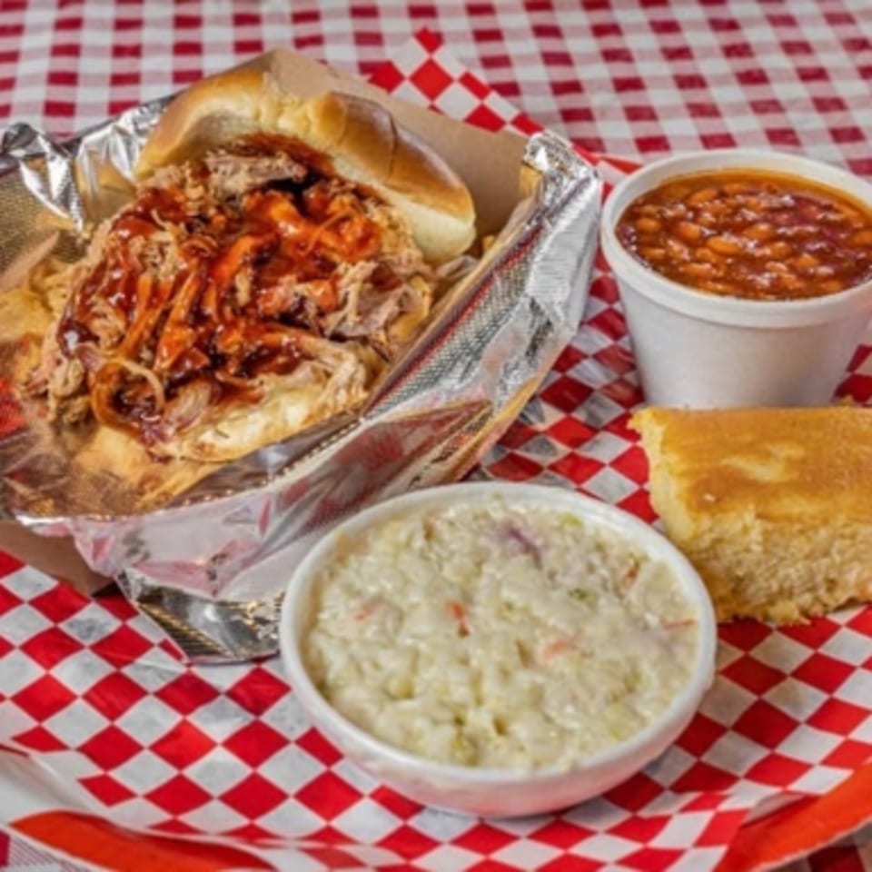 Real Smoq’ed BBQ pulled pork sandwich with baked beans, coleslaw, and cornbread on a checkered tablecloth.