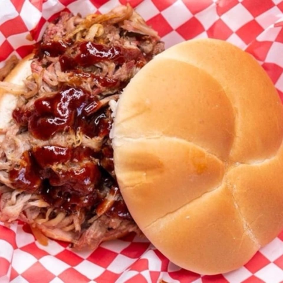 Real Smoq’ed BBQ pulled pork sandwich with barbecue sauce on a bun, in a red and white checkered basket.